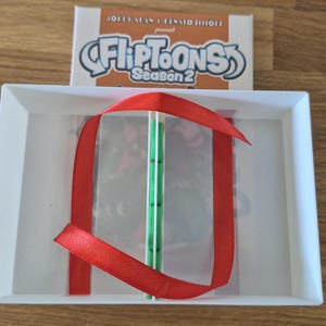 May include: A white rectangular tray containing a green spine book with a red ribbon tied around it. Behind the tray is a white rectangular box with the text "Jordy Alan & Renato Simões present FlipToons Season 2" printed in blue and black.