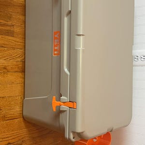 May include: A gray Yeti cooler with orange latches and handles. The cooler is sitting on a wooden floor and has the Yeti logo on the side.