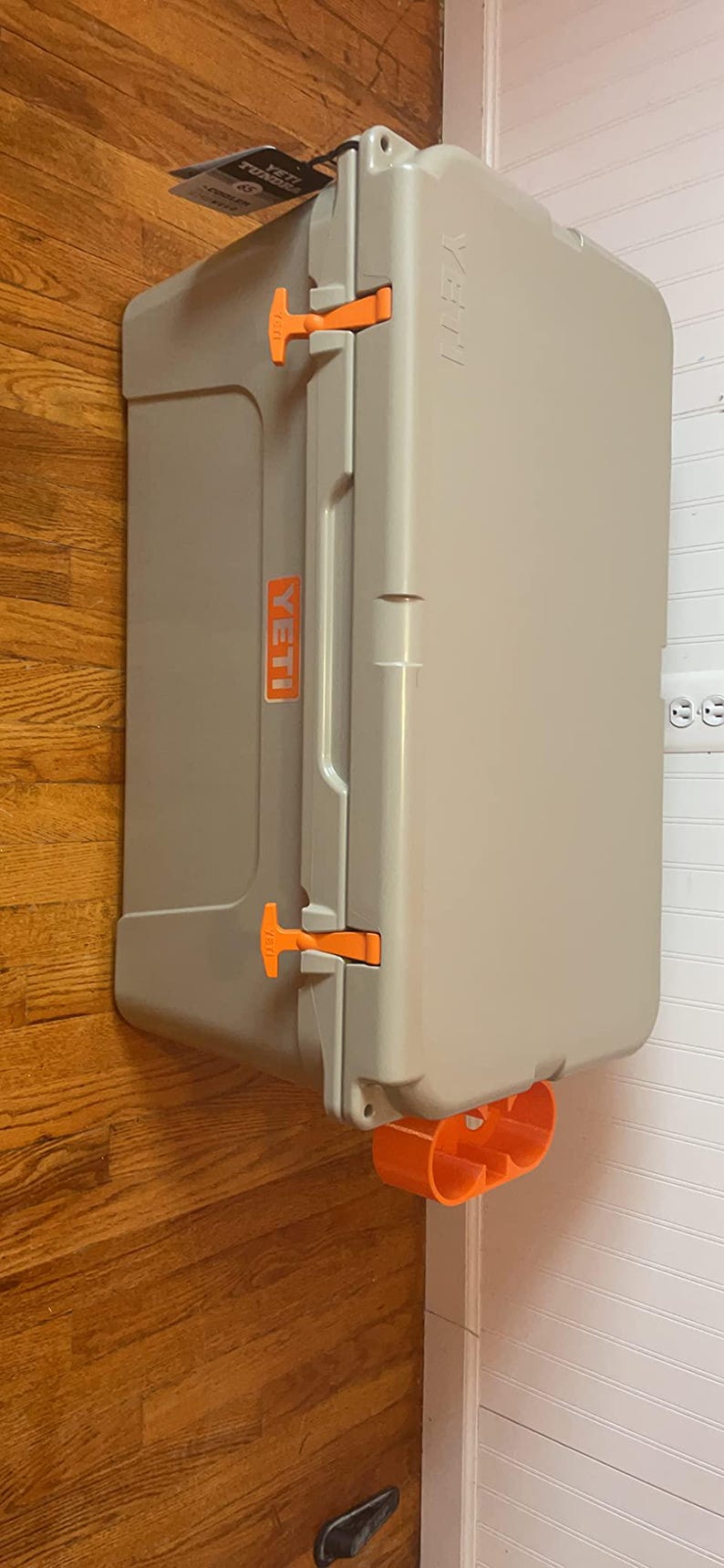 May include: A gray Yeti cooler with orange latches and handles. The cooler is sitting on a wooden floor and has the Yeti logo on the side.