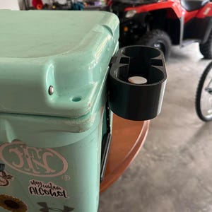 May include: A turquoise cooler with a black cup holder attached to the side. The cooler has a sticker that says "May contain alcohol" and a white Under Armour logo.