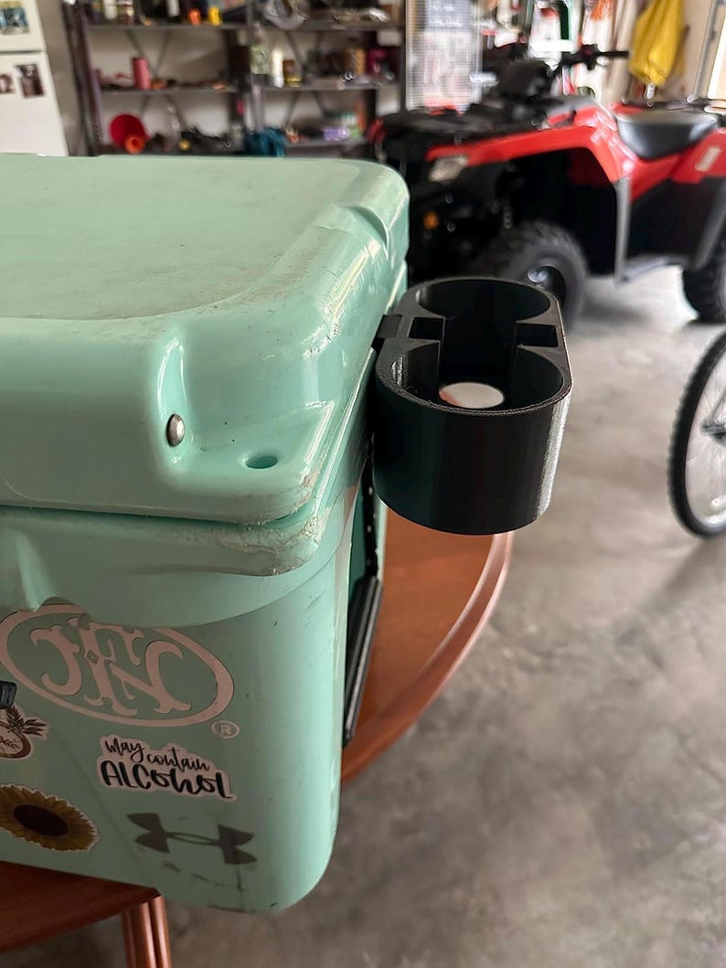 May include: A turquoise cooler with a black cup holder attached to the side. The cooler has a sticker that says "May contain alcohol" and a white Under Armour logo.