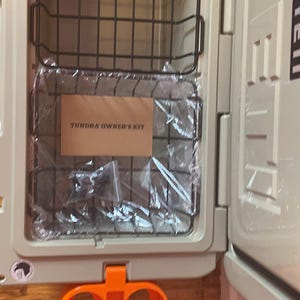 May include: A beige cooler with a black wire basket inside. The cooler has a label that says "Tundra Owner's Kit". The cooler has an orange cup holder on the bottom.