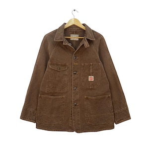 May include: Brown work jacket with four front pockets, button fastenings, and a collared neckline. The jacket is hanging on a wooden hanger. A small patch with the letters "SB" is visible on the front pocket.