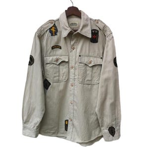 May include: A tan button-down shirt with multiple patches and two chest pockets. The shirt has long sleeves and is hanging on a wooden hanger. Patches include military-style emblems and the word "AIRBORNE".