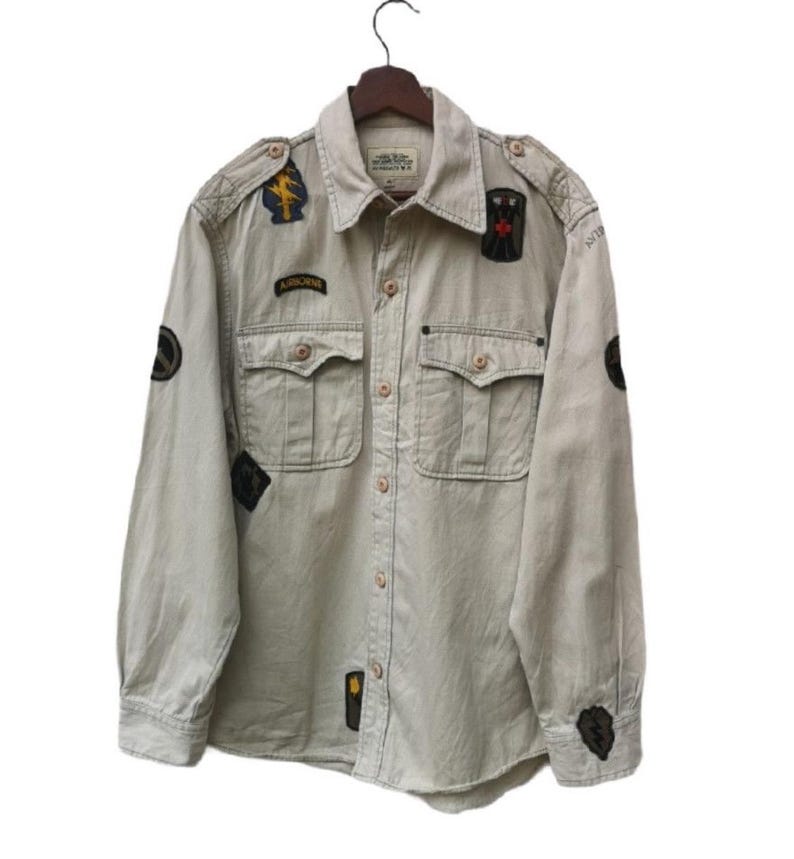 May include: A tan button-down shirt with multiple patches and two chest pockets. The shirt has long sleeves and is hanging on a wooden hanger. Patches include military-style emblems and the word "AIRBORNE".