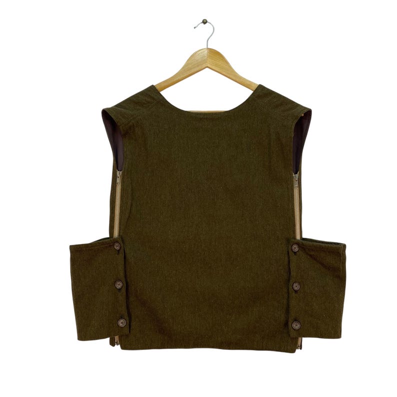 May include: Olive green vest with a round neckline and two side pockets. The vest has a zipper closure on each side and is adorned with brown buttons. The garment is hanging on a wooden hanger against a white background.