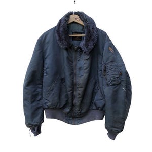 Puede incluir: Chaqueta bomber azul marino vintage con cuello de piel sintética, puños y cintura de canalé. La chaqueta tiene múltiples bolsillos, cierre de cremallera y un pequeño parche en la manga. La chaqueta está colgada en una percha de madera.
