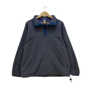 Op de afbeelding: Een grijze fleece trui met een blauwe kraag en accenten. De trui met lange mouwen heeft een knoopsluiting en een klein logo op de borst. De manchetten en zoom zijn ook afgezet met blauw. Het kledingstuk hangt aan een houten hanger.