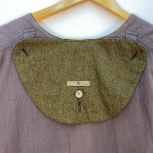 May include: Close-up of a vintage olive green collar with a button and a small rectangular tag with the letter "Y". The collar is attached to a muted purple shirt. The collar has a curved shape and is made of a textured fabric.