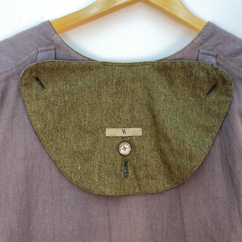 May include: Close-up of a vintage olive green collar with a button and a small rectangular tag with the letter "Y". The collar is attached to a muted purple shirt. The collar has a curved shape and is made of a textured fabric.