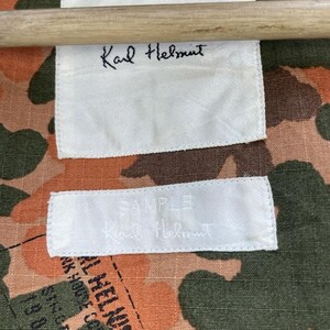 May include: A close-up shot of a camouflage fabric with white labels. The labels display the handwritten name "Karl Helmut" and the word "SAMPLE". The fabric features a blend of orange, brown, and green hues.