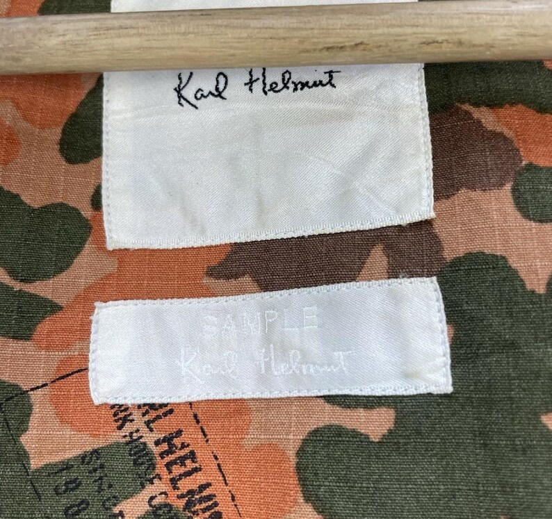 May include: A close-up shot of a camouflage fabric with white labels. The labels display the handwritten name "Karl Helmut" and the word "SAMPLE". The fabric features a blend of orange, brown, and green hues.