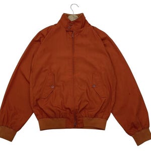 Puede incluir: Una chaqueta bomber de color óxido con cuello, puños y cintura marrones. La chaqueta tiene cierre de cremallera, dos bolsillos delanteros y cuello abotonado. La chaqueta está colgada de una percha metálica.