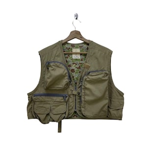 May include: Olive green fishing vest with multiple pockets and a camouflage lining. The vest has a zipper closure and is hanging on a wooden hanger. The vest has the text "Fox Fire" on the inside.