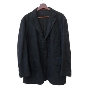 Peut inclure: Un blazer anthracite foncé à fermeture à trois boutons, suspendu à un cintre en bois. Le blazer a un revers cranté et deux poches avant. Le tissu semble être une matière texturée.