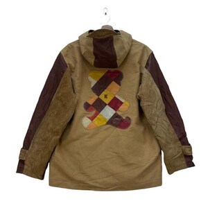 May include: A tan jacket with a brown hood and sleeves. The back features a colourful patchwork teddy bear design. The jacket has a button closure and a hood. The sleeves have a brown leather-like material.