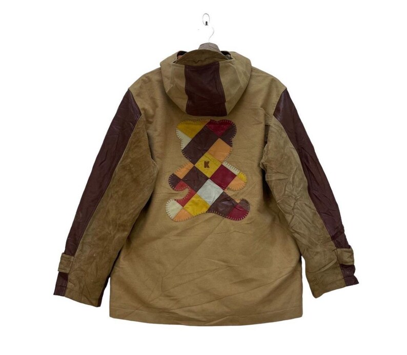 May include: A tan jacket with a brown hood and sleeves. The back features a colourful patchwork teddy bear design. The jacket has a button closure and a hood. The sleeves have a brown leather-like material.