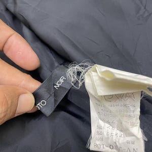 Puede incluir: Una etiqueta de tela negra con el texto "TO NOIR" en letras blancas. Una etiqueta de tela blanca con texto japon&eacute;s e instrucciones de cuidado es visible en el fondo.