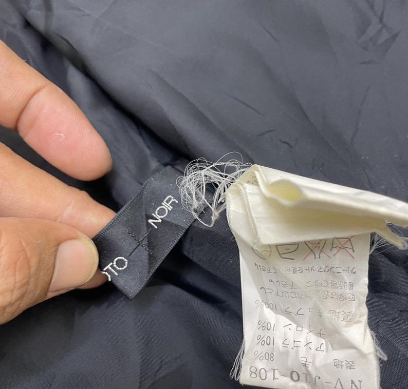 Puede incluir: Una etiqueta de tela negra con el texto "TO NOIR" en letras blancas. Una etiqueta de tela blanca con texto japon&eacute;s e instrucciones de cuidado es visible en el fondo.
