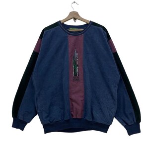 May include: A blue sweatshirt with a maroon vertical stripe down the center and dark green sleeves. The center stripe features an embroidered image of a building. The sweatshirt is hanging on a wooden hanger.