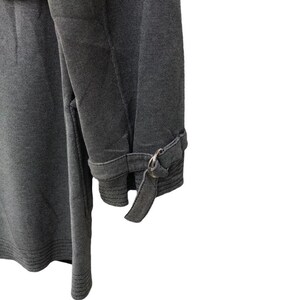 May include: Close-up of a gray fabric garment, possibly a coat or jacket, with a textured appearance. The sleeve features a cuff with a metal ring and a fabric strap detail. The garment is a solid gray color.