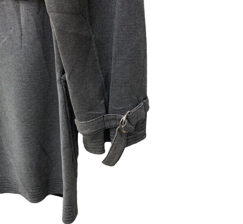 May include: Close-up of a gray fabric garment, possibly a coat or jacket, with a textured appearance. The sleeve features a cuff with a metal ring and a fabric strap detail. The garment is a solid gray color.