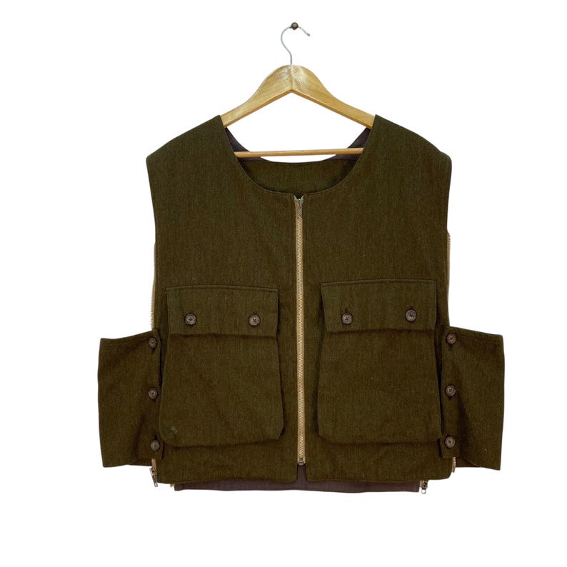 May include: Olive green vest with a full-length zipper and multiple pockets. The vest has four front pockets with button closures and two side pockets. It is hanging on a wooden hanger against a white background.