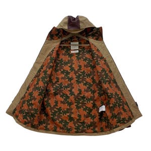 May include: Tan hooded jacket with a brown leather accent on the hood. The interior lining features a camouflage pattern in shades of orange, brown, and green. The jacket is open, revealing the patterned lining and a wooden hanger.