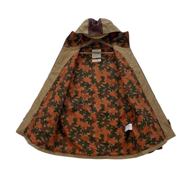 May include: Tan hooded jacket with a brown leather accent on the hood. The interior lining features a camouflage pattern in shades of orange, brown, and green. The jacket is open, revealing the patterned lining and a wooden hanger.