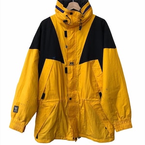 May include: A vintage yellow and black Helly Hansen jacket. The jacket features a high collar with blue accents, a black yoke, and the HH logo. It has multiple pockets and a drawstring waist. The jacket is hanging on a wooden hanger.