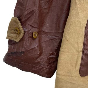 May include: Close-up of a brown leather jacket sleeve with a tan suede panel. The sleeve features a small tan pocket with a button closure and a second button on the leather. The jacket appears to be vintage.