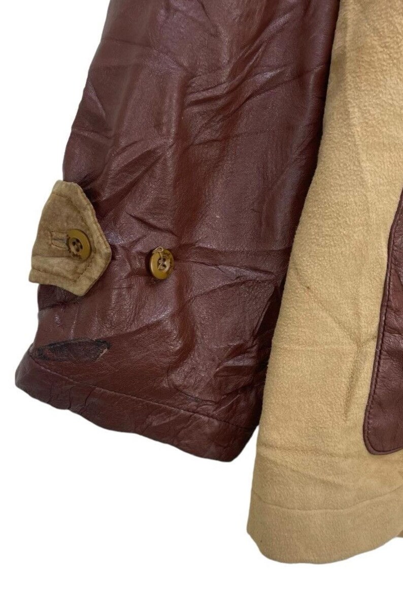 May include: Close-up of a brown leather jacket sleeve with a tan suede panel. The sleeve features a small tan pocket with a button closure and a second button on the leather. The jacket appears to be vintage.