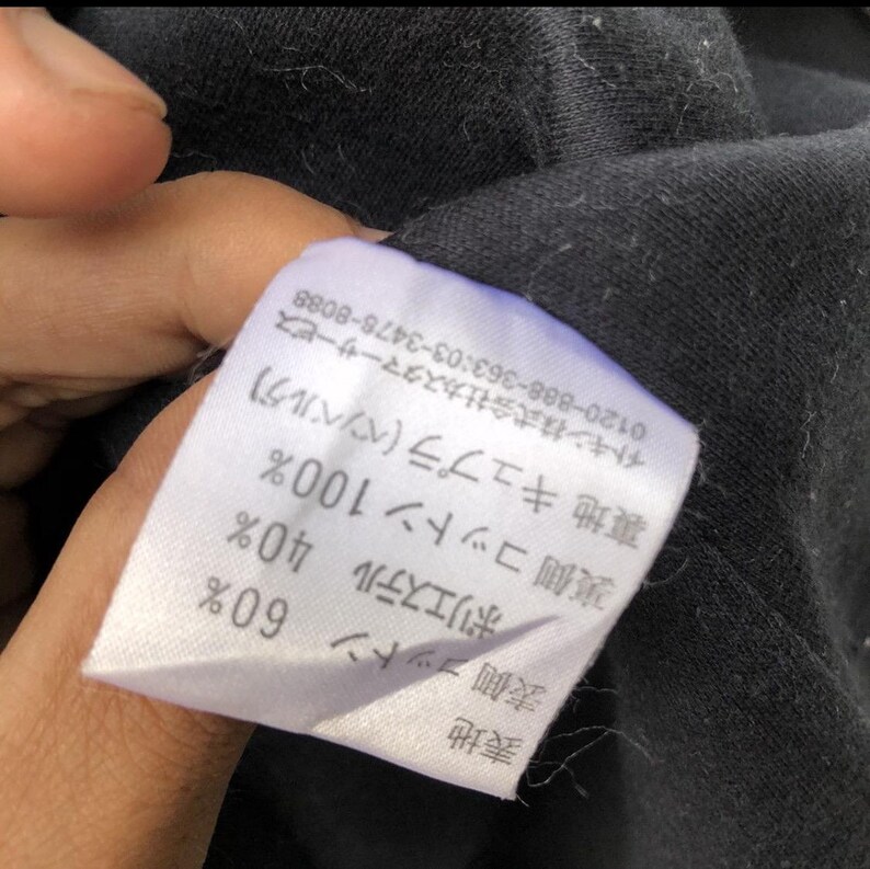 May include: A white fabric tag with Japanese text and percentages for fabric composition. The tag is attached to a black garment. The tag lists the materials as cotton, polyester, and cupro.