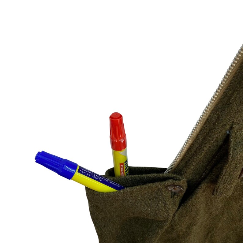 May include: Close-up of a dark olive green fabric with a pocket containing two markers. One marker is blue and yellow, the other is red and yellow. A zipper and button are visible.
