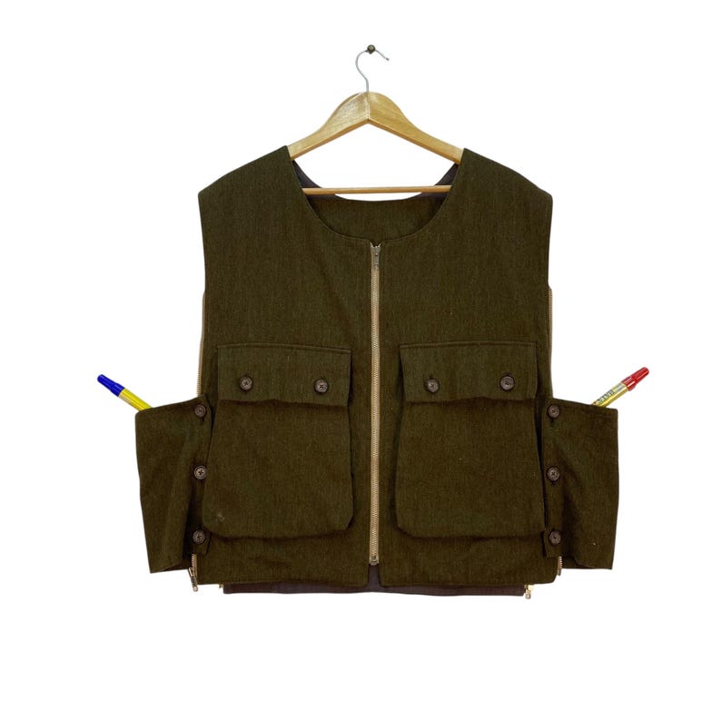 May include: Olive green vest with a full-length zipper, two chest pockets, and two side pockets. The vest has button closures and a brown lining. A blue and yellow pen is in the left pocket, and a red and yellow pen is in the right pocket.