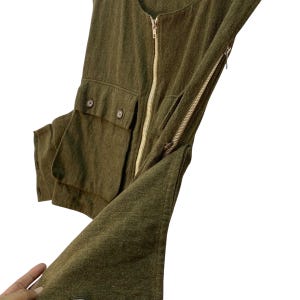 May include: Olive green vest with a full-length gold zipper and two front pockets with button closures. The vest has a textured fabric and a side zipper detail. The garment is shown against a white background.