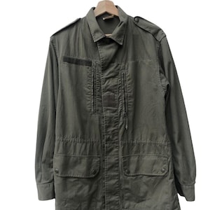 Puede incluir: Chaqueta verde oliva con cuello, charreteras y múltiples bolsillos. La chaqueta tiene una botonadura frontal y dos bolsillos en el pecho con cremallera. Cuelga de una percha de madera.