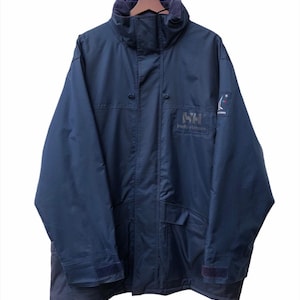 May include: Navy blue Helly Hansen jacket with a high collar and multiple pockets. The jacket features the Helly Hansen logo on the chest and a small emblem on the sleeve. The jacket is hanging on a wooden hanger.