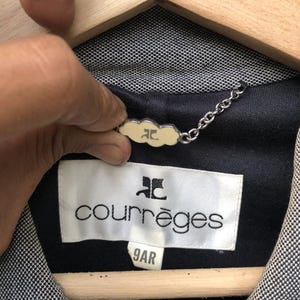 May include: Close-up of a Courr&egrave;ges jacket label. The label is white with black text and a logo. The jacket is black and white with a small cream-colored tag attached by a chain.