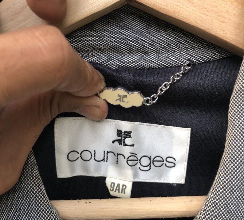 May include: Close-up of a Courr&egrave;ges jacket label. The label is white with black text and a logo. The jacket is black and white with a small cream-colored tag attached by a chain.