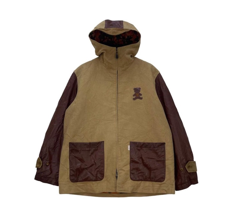 May include: A tan jacket with a hood and brown sleeves. The jacket features two front pockets and a small teddy bear patch on the chest. The hood lining has a floral pattern.