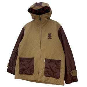 May include: Tan hooded jacket with brown leather sleeves and pocket details. Features a small teddy bear patch on the chest. The hood has a brown leather accent and a patterned lining. Includes a full-length zip and two front pockets.