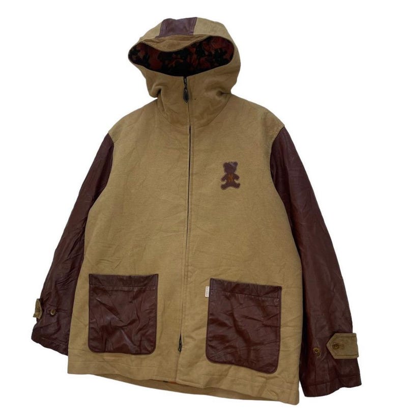 May include: Tan hooded jacket with brown leather sleeves and pocket details. Features a small teddy bear patch on the chest. The hood has a brown leather accent and a patterned lining. Includes a full-length zip and two front pockets.