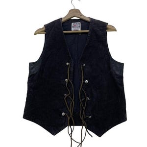 Może przedstawiać: Bordowa kamizelka zamszowa z elementami skórzanymi i brązowymi skórzanymi sznurówkami. Kamizelka ma szpiczasty kołnierz i zapięcie z przodu na metalowe zatrzaski.