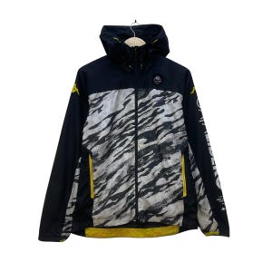 Op de afbeelding: Zwart-witte jas met capuchon en een zebra streep patroon op de voorkant. De jas heeft een zwarte capuchon, mouwen en rug, met gele accenten op de rits en het logo. De jas hangt aan een houten hanger.