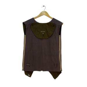 May include: A sleeveless vest in shades of brown and olive green, hanging on a wooden hanger. The vest features a large olive green pocket on the back, two side zippers, and adjustable straps with buckles. The vest is made of a durable fabric.