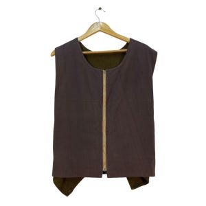 May include: A reversible vest with a brown exterior and a green interior, hanging on a wooden hanger. The vest has a full-length zipper down the front.