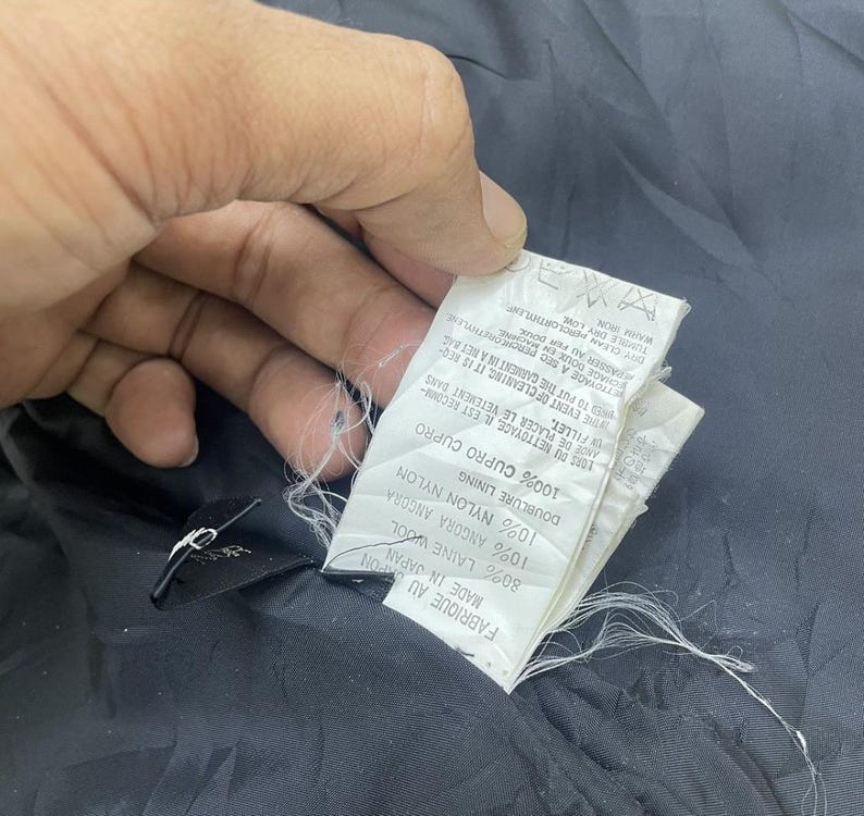 Puede incluir: Una etiqueta de tela negra con texto blanco que detalla la composici&oacute;n de la tela de la prenda. La etiqueta dice "Fabrique au Japan, Made in Japan, 80% Laine Wool, 10% Angora Angora, Doublure Lining, 10% Nylon Nylon, 100% Cupro Cupro".