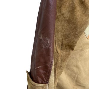 May include: Close-up of a vintage jacket with a brown leather sleeve and tan suede body. The jacket has a pocket and a patch detail. The leather sleeve shows signs of wear.