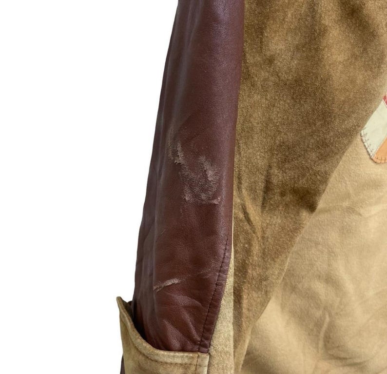 May include: Close-up of a vintage jacket with a brown leather sleeve and tan suede body. The jacket has a pocket and a patch detail. The leather sleeve shows signs of wear.